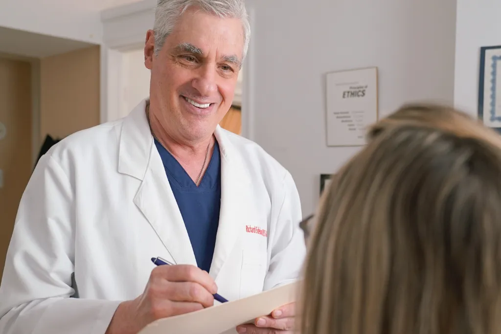 A young adult in a modern dental chair as the dentist, in white lab coat, gently explains a procedure. The sunlit, organized clinic feels welcoming and reassuring.